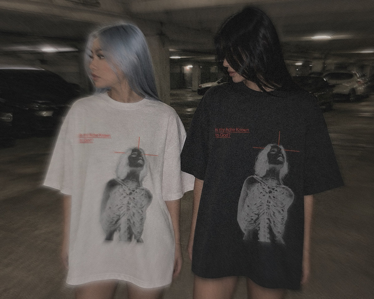 Two models wearing oversized graphic t-shirts in a dimly lit parking garage. The model on the left has light blue hair and wears a white t-shirt with a black and white graphic of a skeletal figure under a red cross, with small red text.