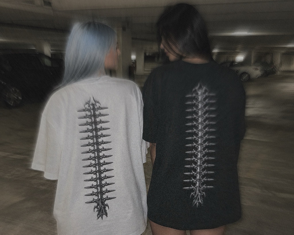 Two people seen from behind in a dimly lit parking garage, wearing oversized T-shirts with large vertical vertebrae with ammo prints on the back. One shirt is off-white and the other is black.