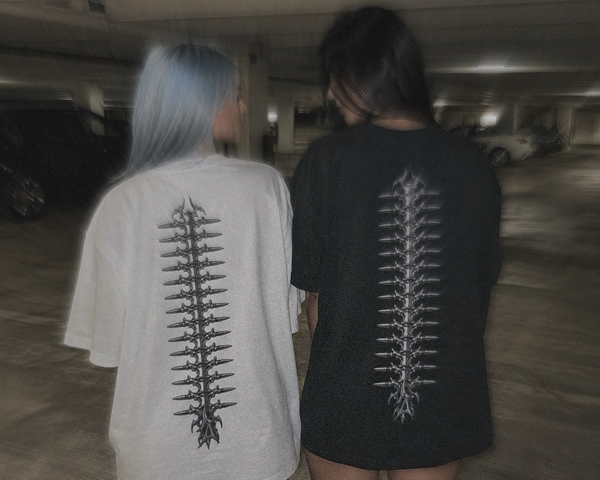 Two people seen from behind in a dimly lit parking garage, wearing oversized T-shirts with large vertical vertebrae with ammo prints on the back. One shirt is off-white and the other is black.
