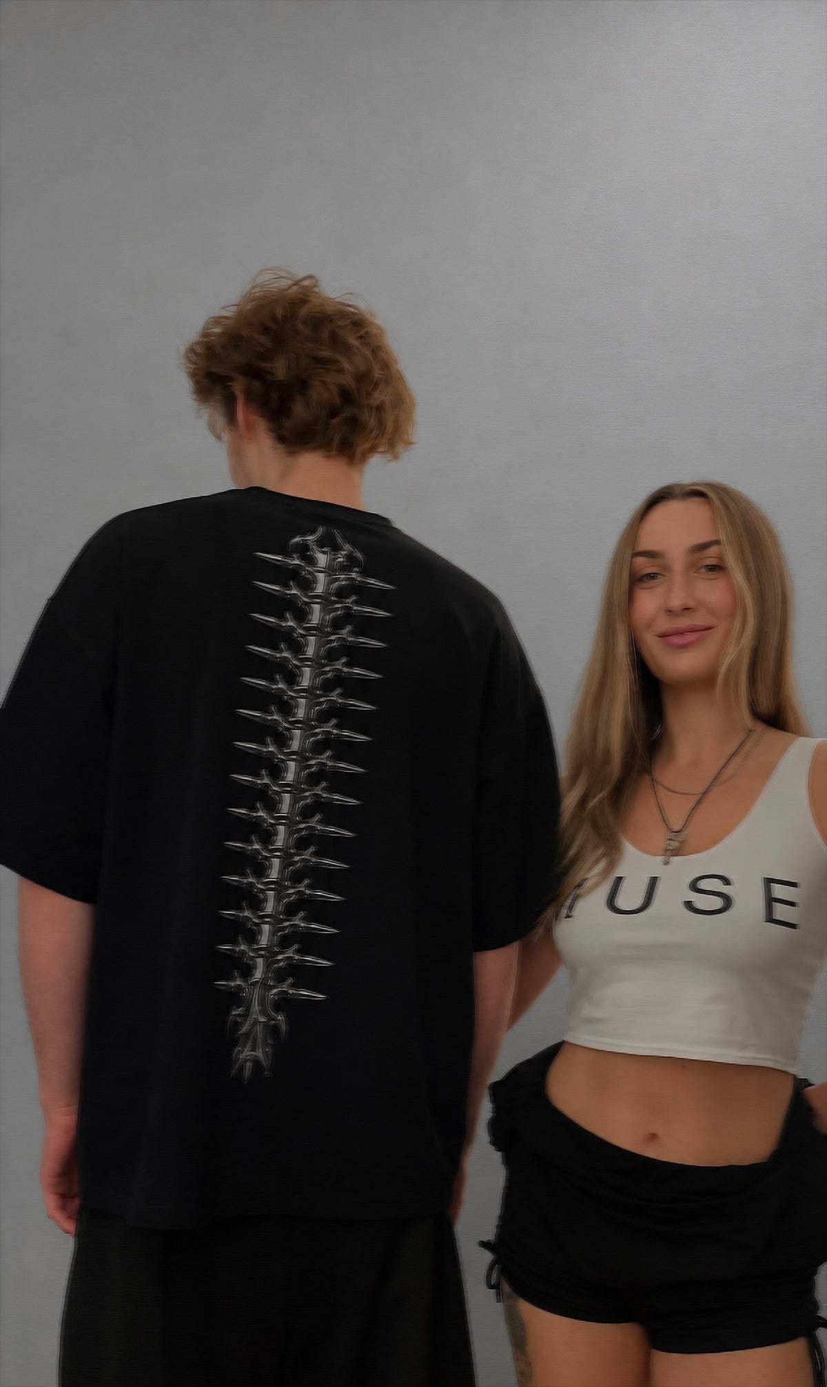 A man wearing a black oversized t-shirt with a large white spine graphic on the back, standing next to a woman wearing a white crop top with 'MUSE' printed on it.