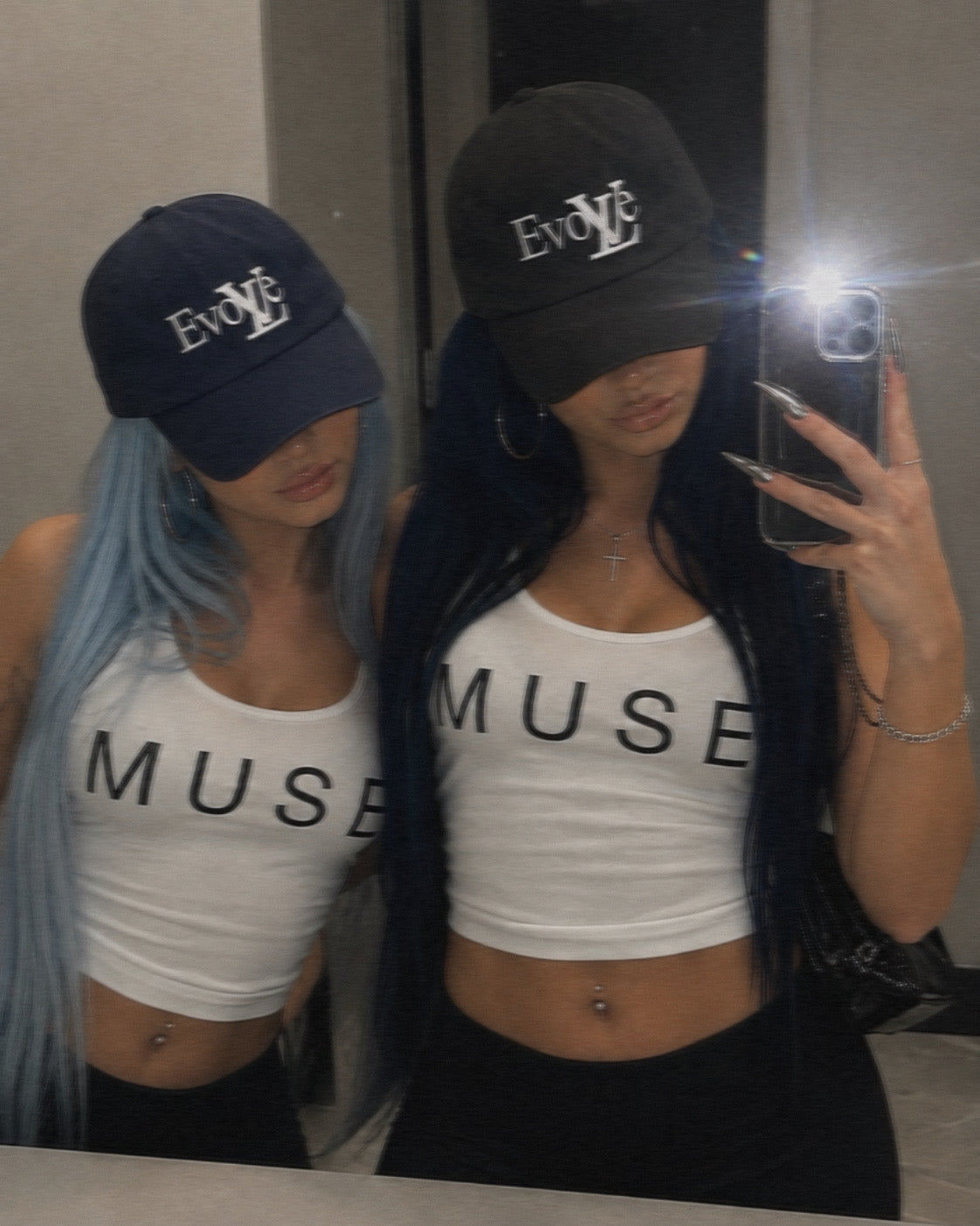 Two women taking a mirror selfie wearing matching outfits: white cropped tank tops with black 'MUSE' text and black baseball caps with white 'EvOLVE' text. One woman has long light blue hair and the other has long dark hair.