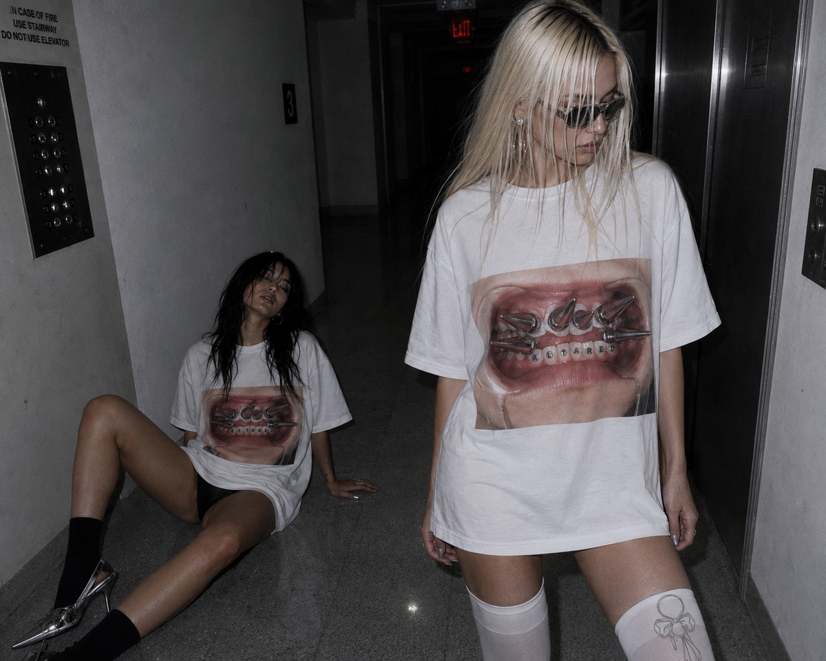 Two women in a dimly lit hallway wearing oversized white graphic T-shirts featuring a large, detailed photo print of TEETH WITH THE WORD ALTARED SPELLED OUT WITH TOOTH GEMS on the front. One woman stands while wearing the shirt and sunglasses, and the other sits on the floor nearby.