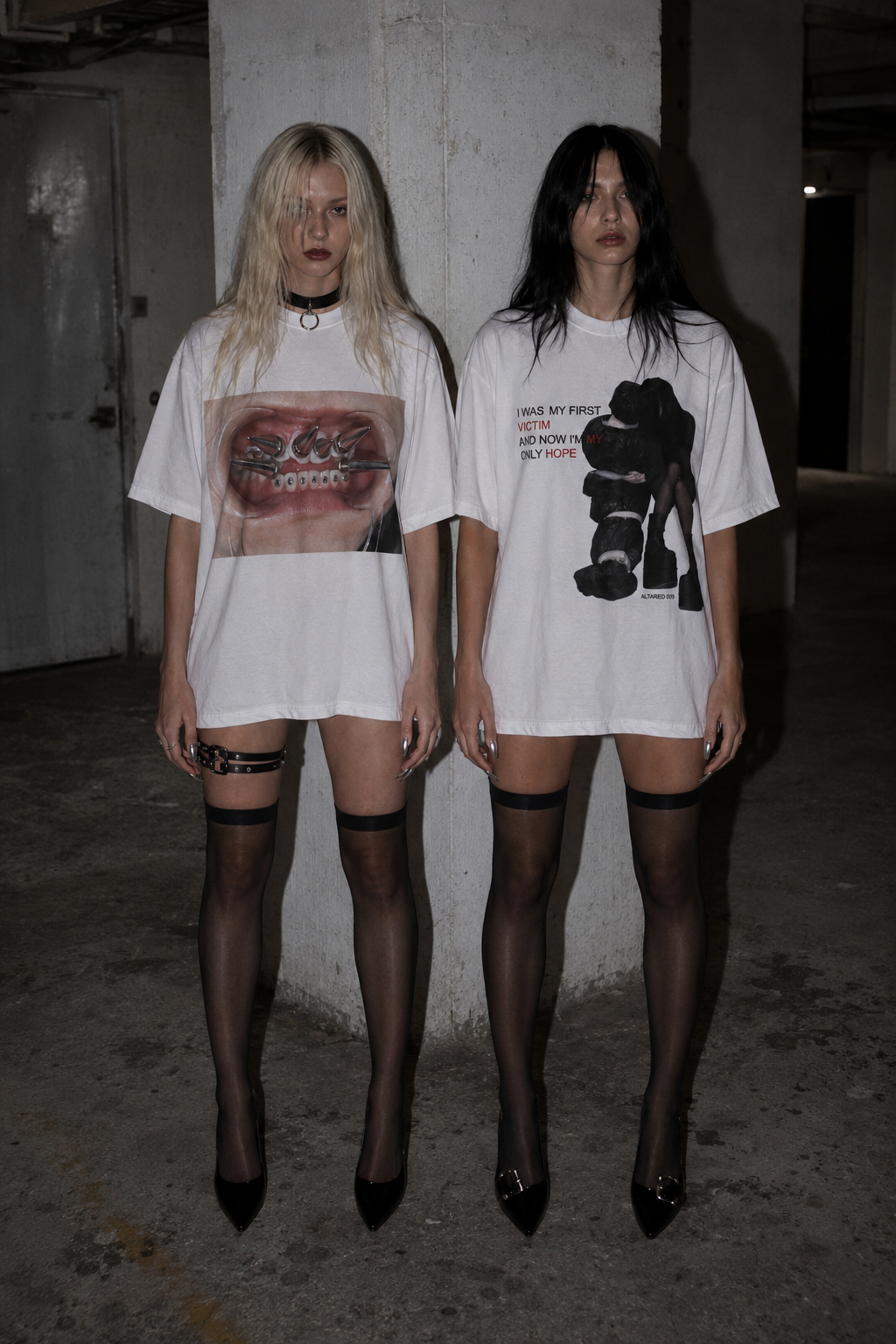 Two women standing in a dimly lit concrete parking structure wearing oversized white graphic T-shirts, black thigh-high stockings, and black heels.