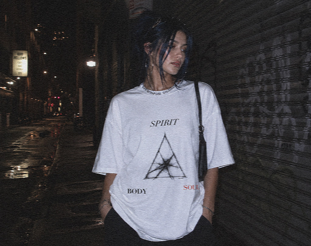 A person standing on a dark city street at night wearing an oversized white graphic T-shirt with the word 'SPIRIT' above a geometric symbol and 'BODY SOUL' text, paired with black bottoms and a black shoulder bag.